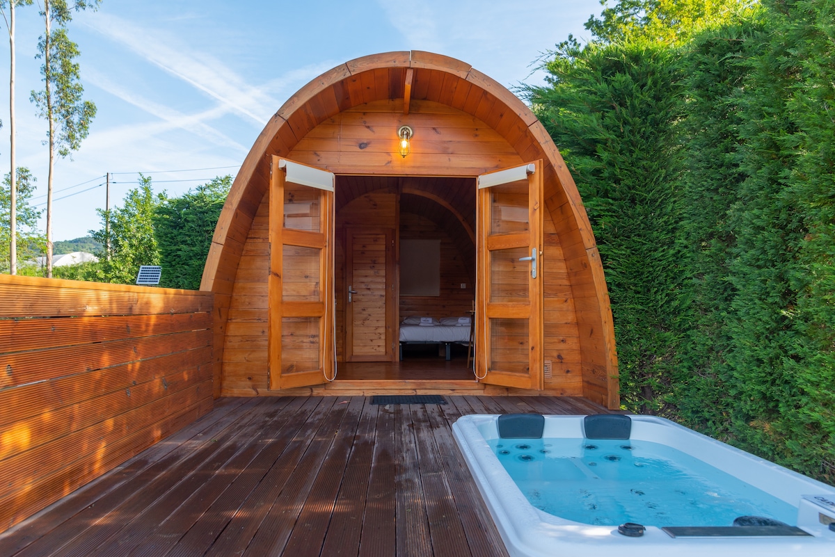 A wooden structure featuring an arched design is highlighted by open double doors, inviting natural light. A private hot tub is positioned on the deck, providing an area for relaxation. Lush greenery surrounds the space, enhancing its connection to nature.