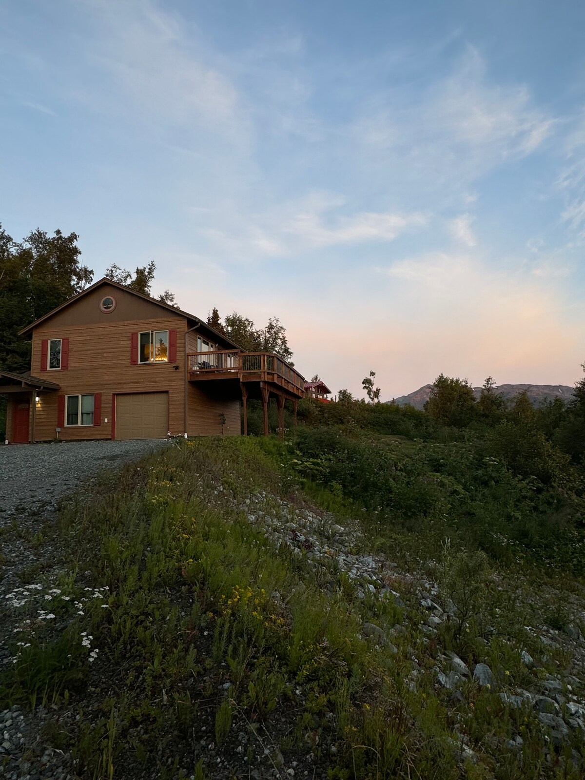 A mountain cabin is set against a sky painted with soft pastel hues. The structure features two stories, a deck extending from the upper level, and is surrounded by greenery. Wildflowers bloom in the foreground, adding natural beauty to the serene landscape.