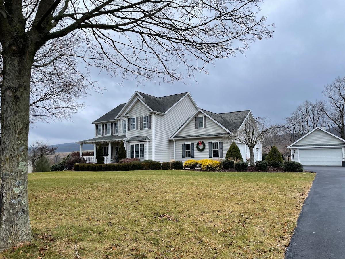 A charming two-story home is set on a grassy lawn, surrounded by neatly trimmed hedges and small shrubs. A large front porch provides an inviting entry, while a detached garage is located to the right. The sky is overcast, hinting at a serene atmosphere.