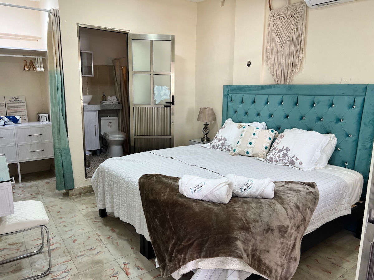 A well-lit sleeping area is featured, highlighted by a spacious bed with a tufted turquoise headboard. Soft linens and throw blankets in neutral tones are arranged at the foot of the bed. A doorway leads to a separate bathroom area, enhancing the overall functionality of the space.