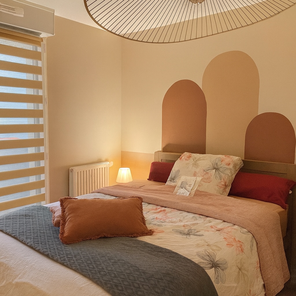The private bedroom features a spacious bed adorned with a floral-patterned quilt, complemented by textured pillows in warm tones. A side lamp provides soft lighting, while a patterned ceiling fixture adds an artistic touch. The window is dressed with sheer blinds, allowing natural light to filter in.