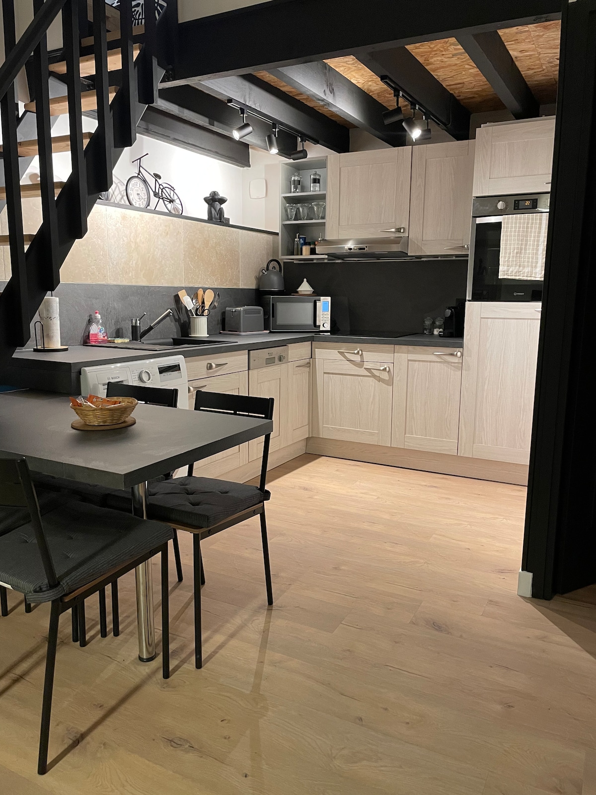 A modern kitchen area features light-colored cabinetry and essential appliances, including a microwave and dishwasher. A dining table with four black chairs sits on the polished wooden floor, complemented by tasteful kitchenware. Soft lighting highlights the organized space, enhancing its functional appeal.