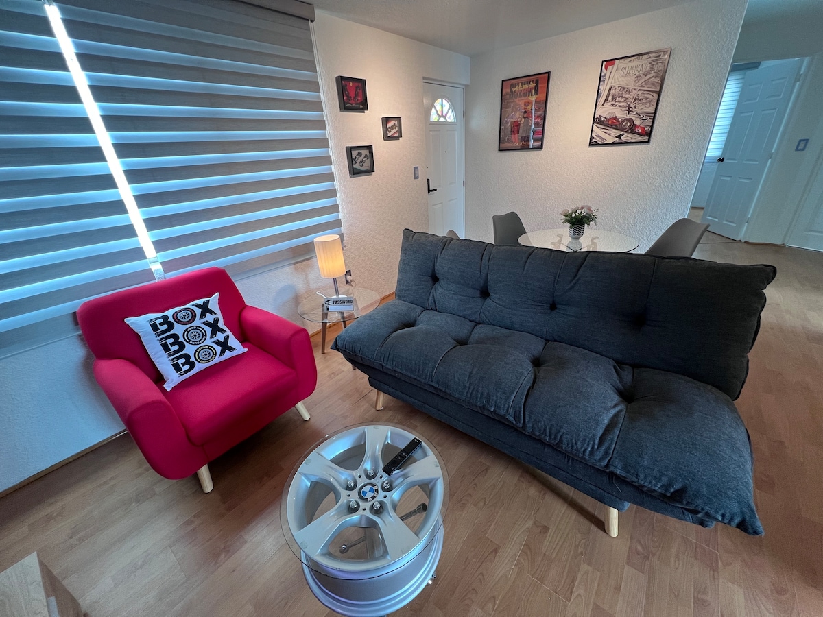 A modern living area is presented with a dark gray futon and a vibrant red armchair featuring a decorative cushion. A round table is set for dining, accompanied by four chairs. Natural light enters through window treatments, highlighting the wood flooring.