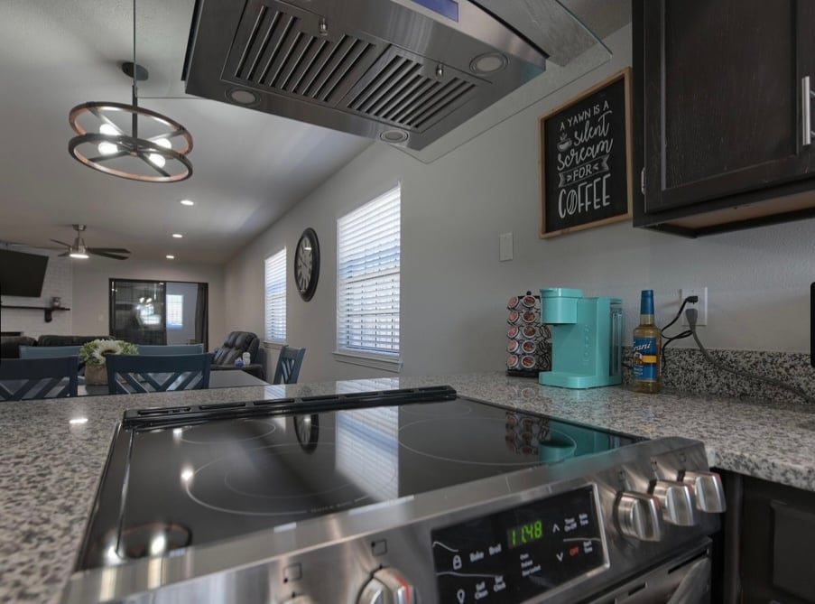 A modern kitchen is showcased, featuring a sleek stovetop with a stainless steel finish and a contemporary ventilation hood above. Granite countertops are visible, accompanied by a turquoise coffee maker and a decorative chalkboard sign that reads about the joy of coffee.
