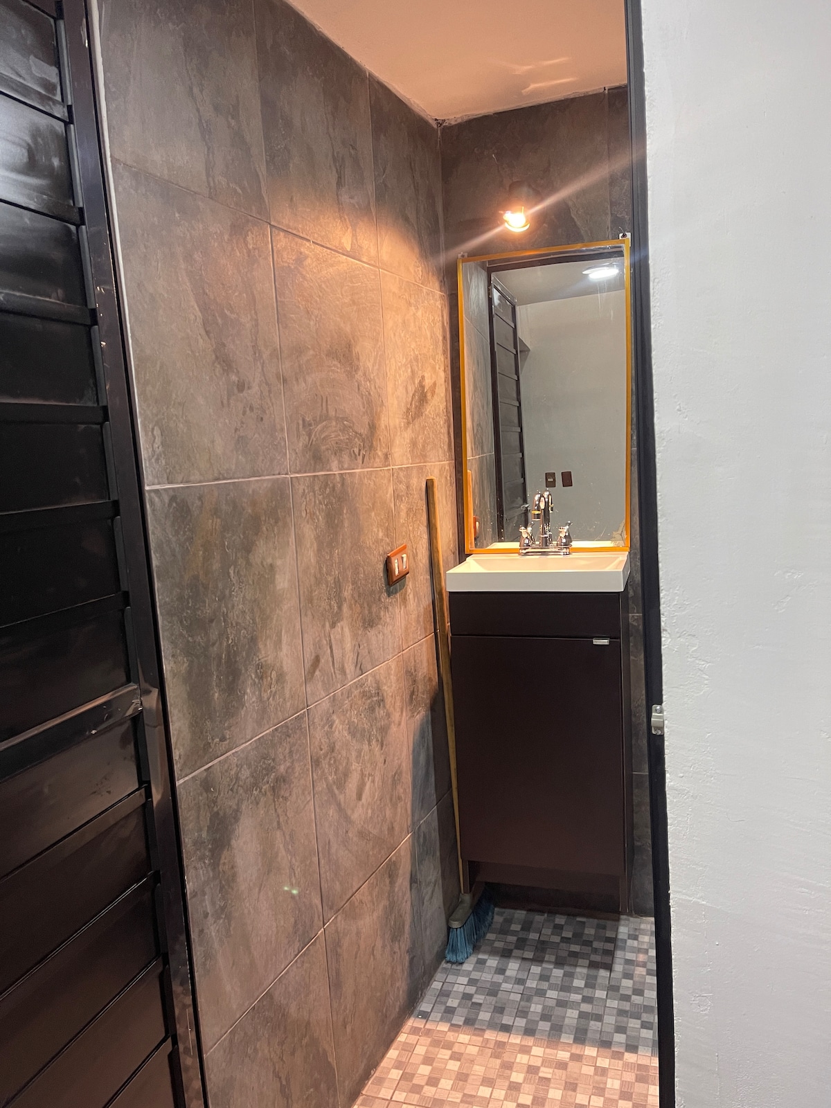 A compact bathroom space features textured gray walls and a modern sink with a stylized mirror above it. The lighting is provided by a wall-mounted fixture, while a patterned floor adds an element of interest to the room.