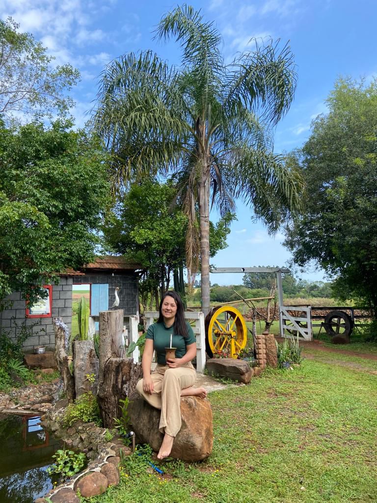 A large stone serves as a natural seat under the shade of a tall palm tree. A rustic building is visible in the background, accented by colorful elements. Lush greenery surrounds the area, creating a serene atmosphere in an outdoor setting.