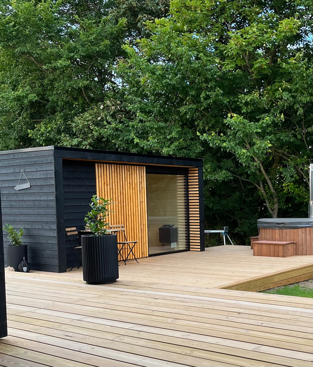 A modern exterior of a compact building showcases a wooden deck surrounded by trees. A large glass door with wooden slats offers a glimpse inside, while potted plants and a small table with chairs provide seating. A wood-fired hot tub is visible on the right.