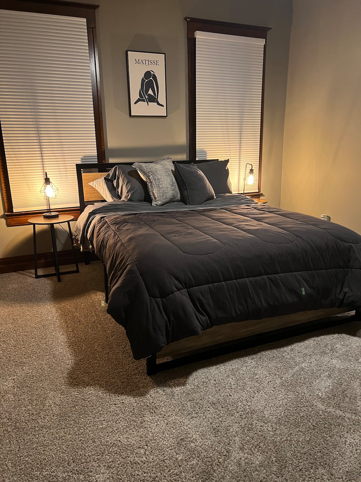 A sleeping area features a dark bedding set atop a spacious bed, complemented by bedside lamps providing soft illumination. Two windows with blinds allow natural light in, while a framed art piece enhances the wall above the bed. Plush carpeting adds warmth to the room.