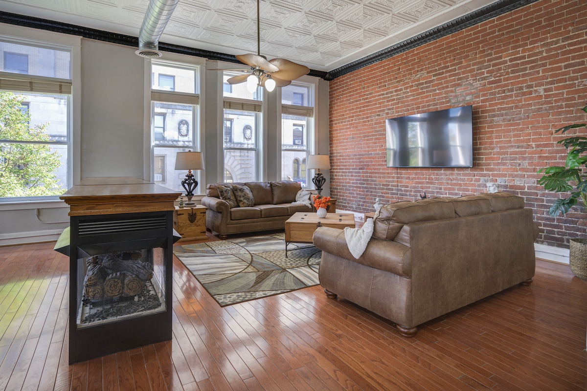An inviting living area features exposed brick walls and large windows allowing natural light. Two comfortable sofas are arranged around a decorative area rug, with a wooden table nearby. A flat-screen TV is mounted on the wall, and a ceiling fan circulates air.