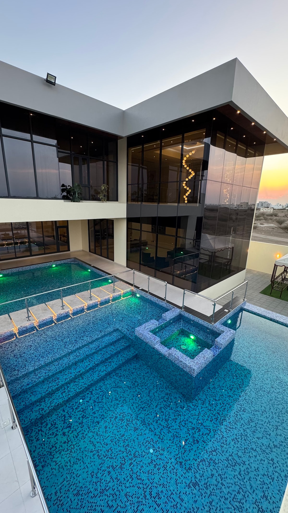 A modern building is showcased with an expansive pool featuring steps leading into the water. The pool area is framed by sleek glass walls, reflecting the evening light. Green lighting enhances the design of the pool, and an elevated seating area is visible along the side.
