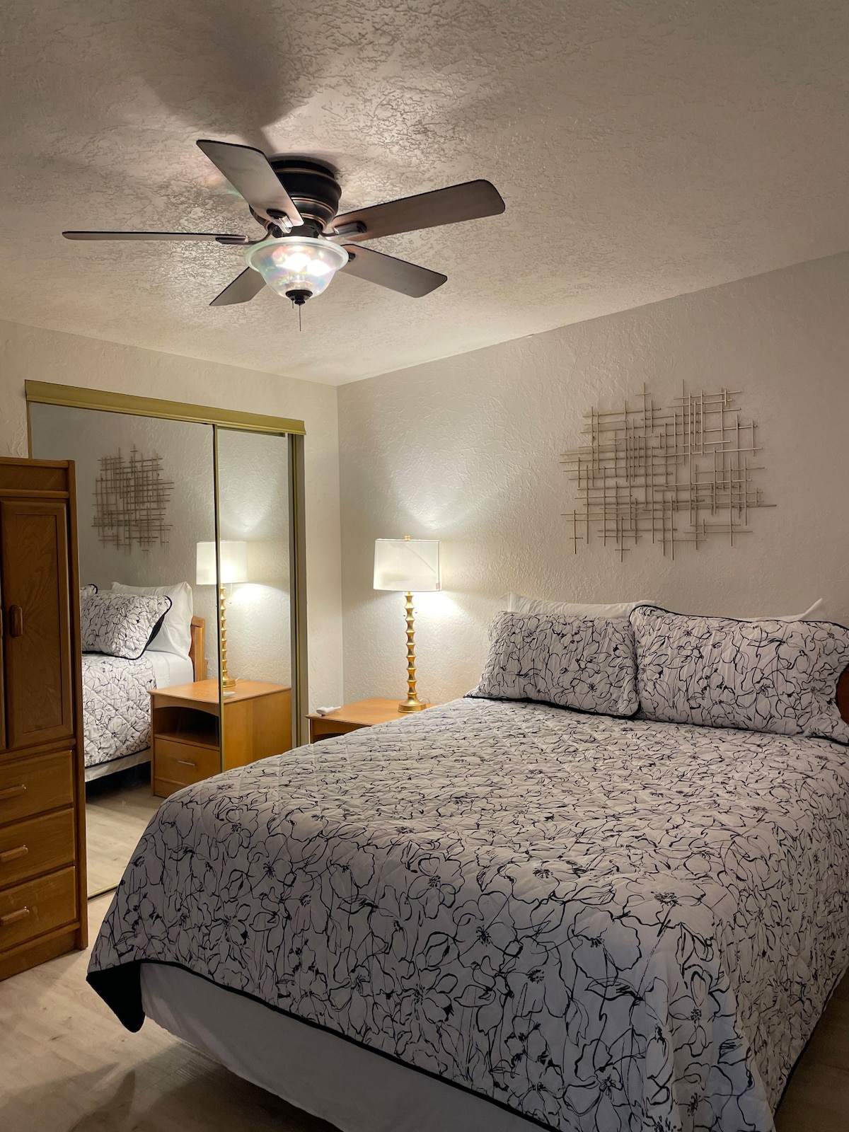A well-lit bedroom features a spacious bed adorned with a floral-patterned duvet. A bedside lamp sits next to the bed, providing soft illumination. A mirror reflects the room, enhancing its openness, while a ceiling fan adds comfort and style to the space.