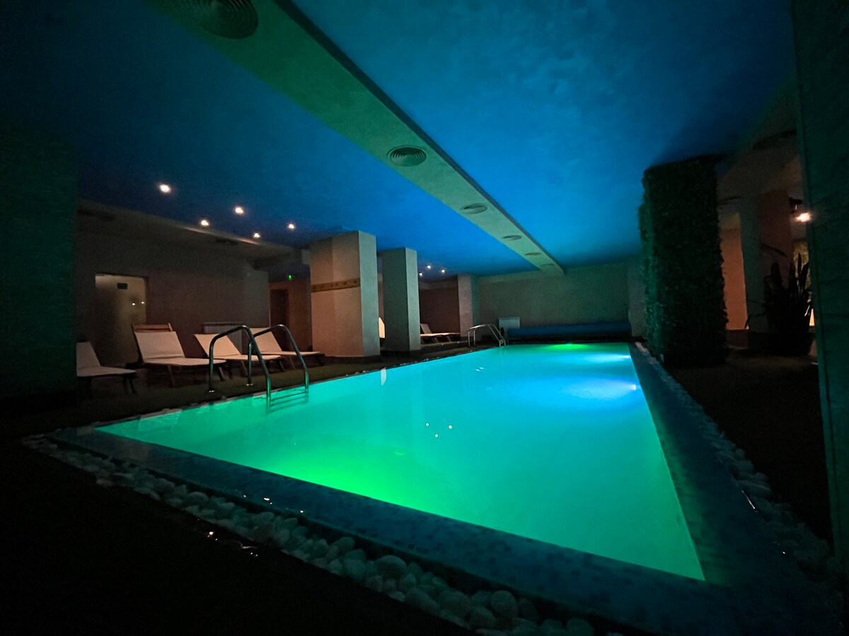 An indoor pool is illuminated with varying shades of blue and green light, creating a serene ambiance. Comfortable lounge chairs line the edges, and smooth stones are arranged around the pool's perimeter. The dim lighting enhances a relaxing atmosphere.