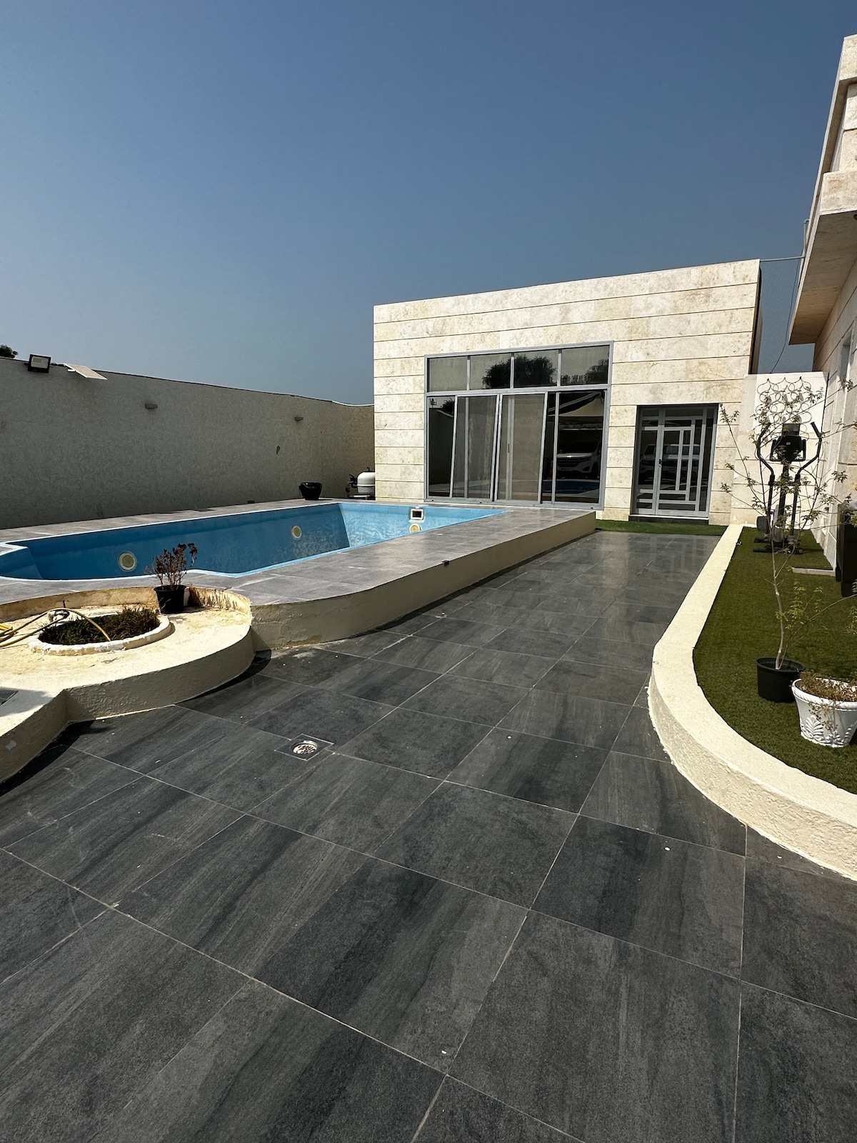 An outdoor area features a swimming pool surrounded by a sleek stone patio. Large glass doors offer a view of the interior. Planters with greenery are positioned along the edge, contributing to a serene environment under a clear blue sky.