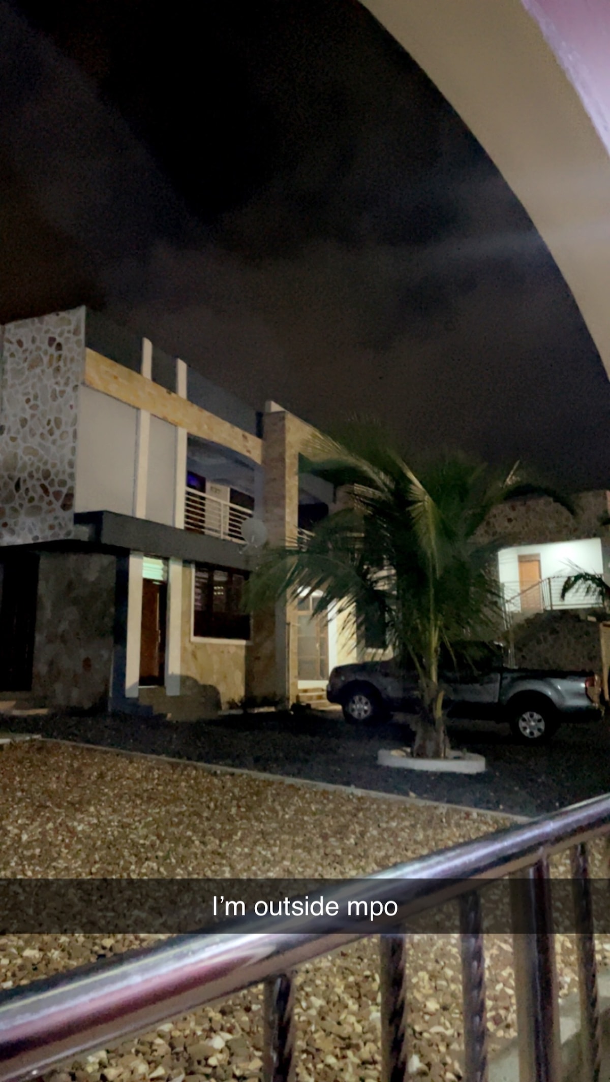 The exterior of a multi-story building is visible at night, featuring a combination of stone and modern materials. A palm tree is located near the entrance, and a vehicle is parked in view. Soft lighting highlights architectural details.