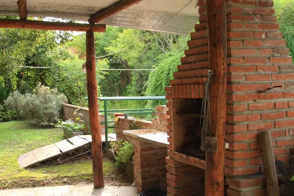 An outdoor grilling area constructed with rustic brick features a barbecue grill, surrounded by greenery and natural elements. A wooden patio covers part of the space, providing shade. The area overlooks a lush garden, contributing to a serene outdoor environment.