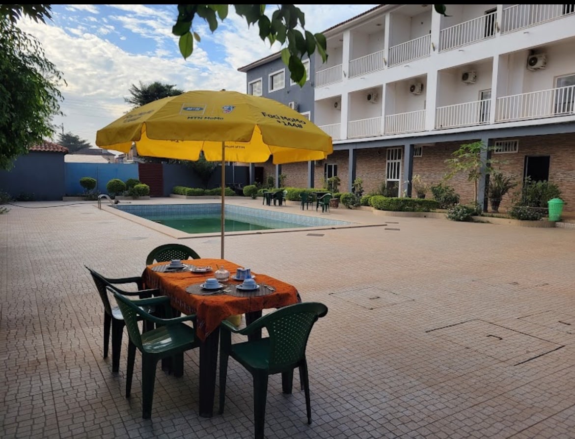An outdoor dining area with a table set for two is featured under a yellow umbrella. Nearby, an inviting rectangular pool reflects the sky, while a surrounding patio is paved with a light-colored tile. Lush greenery and shrubs enhance the space's charm.
