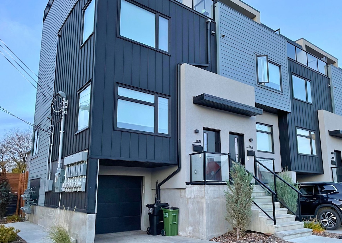 The modern townhome features a sleek exterior with a mix of gray and black cladding. Large windows provide natural light, while a glass railing leads to the entrance. A garage is visible at the base, complemented by well-maintained landscaping.