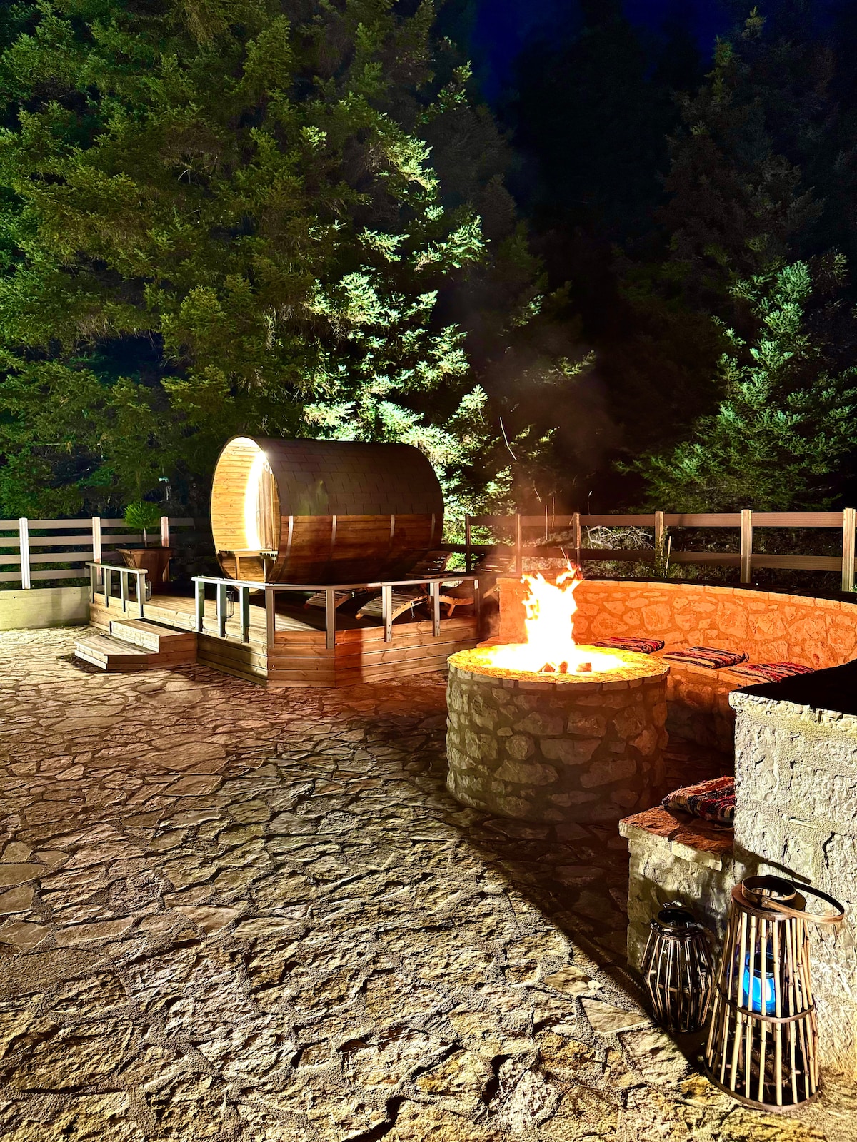 An outdoor area features a wooden sauna adjacent to a stone fire pit, both surrounded by forested scenery. The sauna, crafted from natural materials, complements the stonework of the fire pit, which is currently ablaze. The setting is illuminated by the firelight, creating a cozy ambiance.