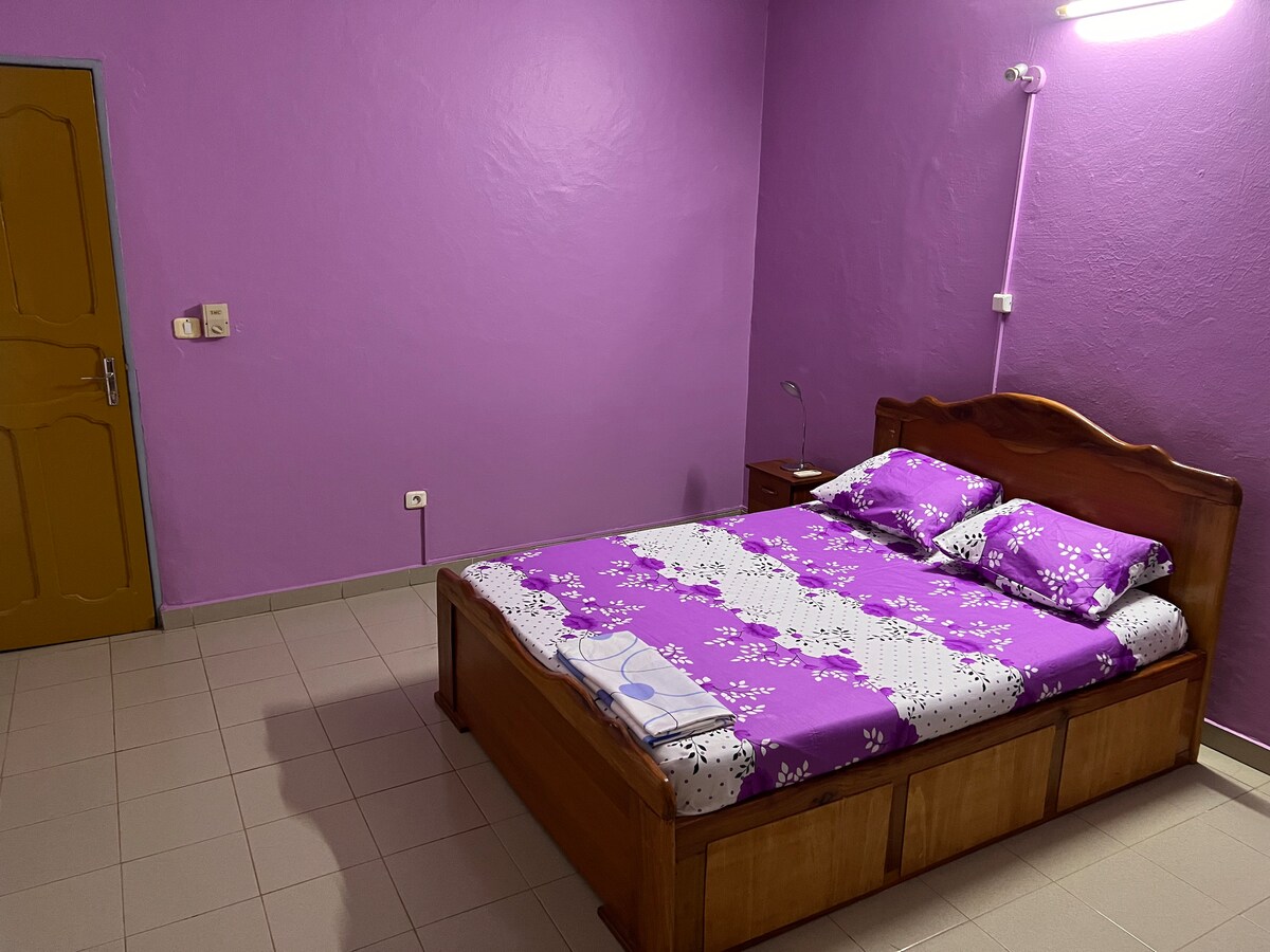 A sleeping area features a wooden bed with a floral-patterned purple bedspread, complemented by matching pillowcases. The room is painted in a solid purple hue, and a bedside table with a lamp is positioned to the right of the bed. Light-colored tiles cover the floor.