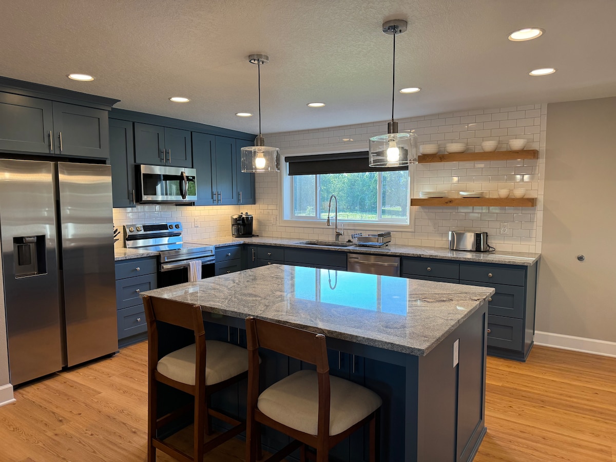 A modern kitchen is shown with dark blue cabinetry and a spacious island topped with light-colored granite. Two wooden stools are positioned at the island. Stainless steel appliances are integrated, and shelves above the counter display neatly arranged dishware.