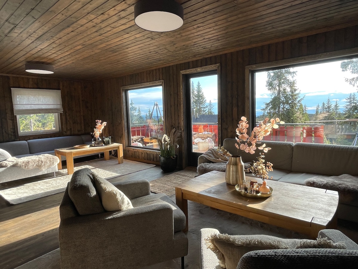 A spacious living area features large windows offering scenic views of surrounding nature. Comfortable seating is arranged around wooden coffee tables, and decorative accents, including flowers, enhance the atmosphere. A mix of textures and materials is evident in the furniture and decor.