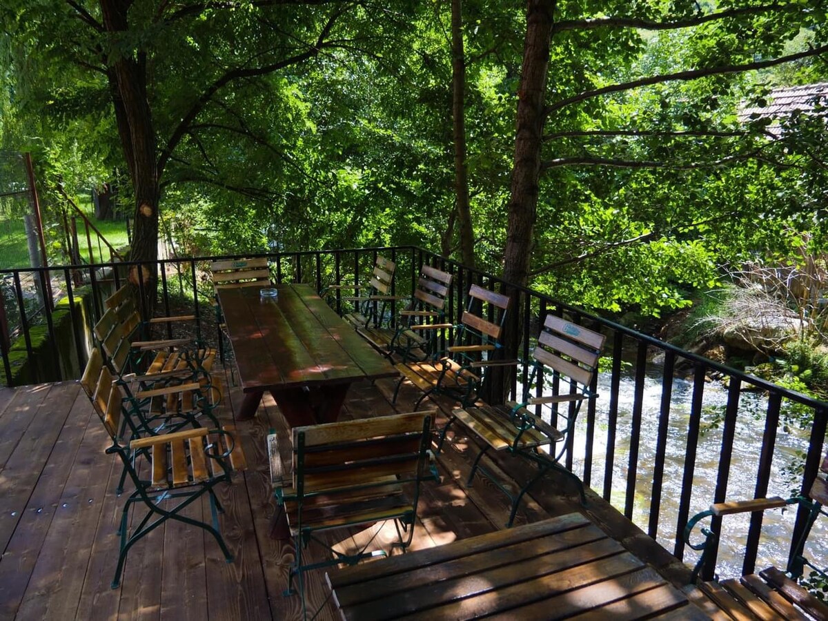 An outdoor dining area is surrounded by lush greenery, featuring a long wooden table with eight matching chairs. A gentle stream flows nearby, creating a serene backdrop. The deck's wooden flooring complements the natural setting.