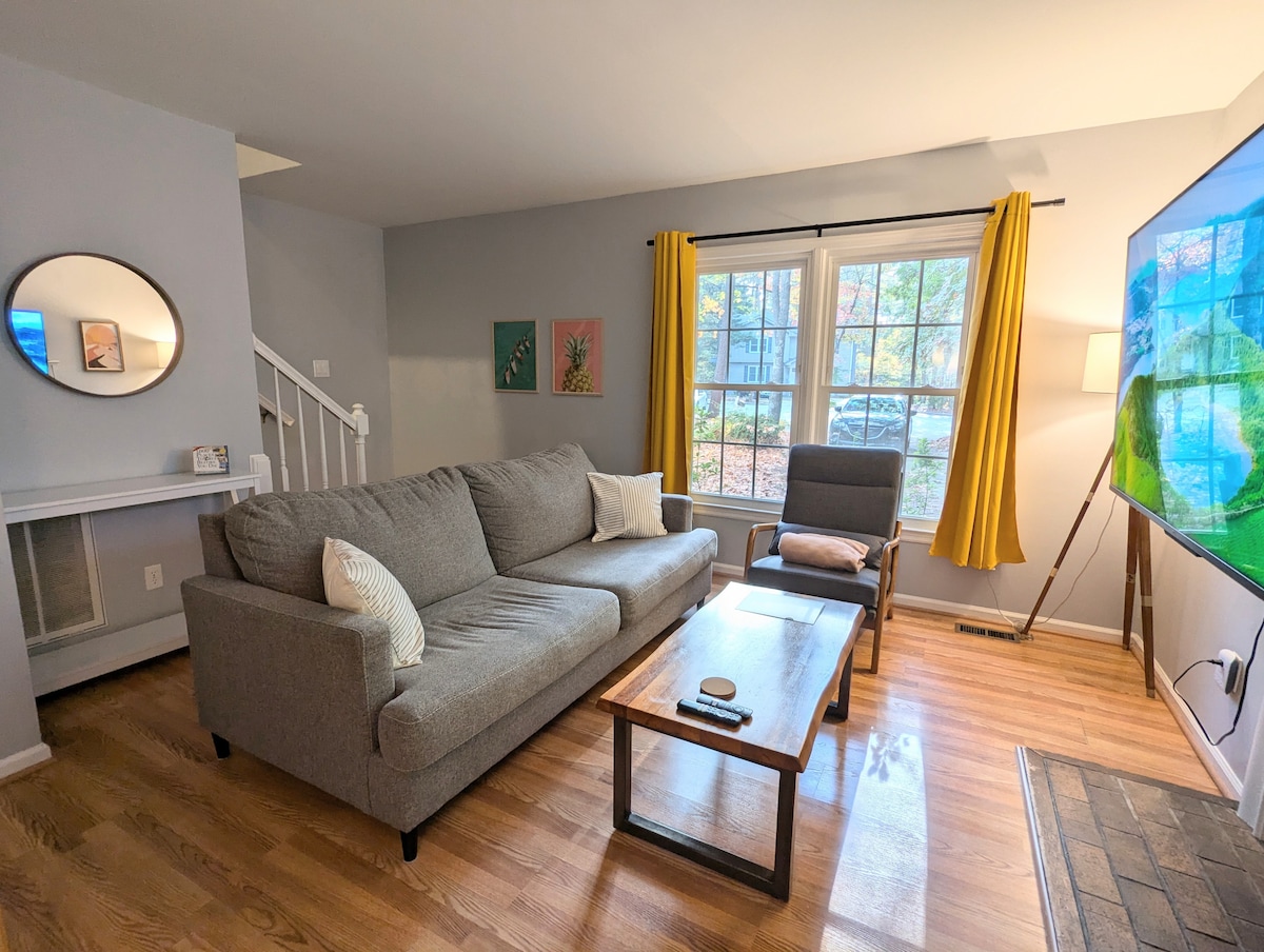 A living area features a comfortable gray sectional sofa with decorative pillows, paired with a gray armchair. A coffee table sits in front, and a large television is mounted on the wall. Natural light filters through the window, adorned with yellow curtains, creating a bright atmosphere.