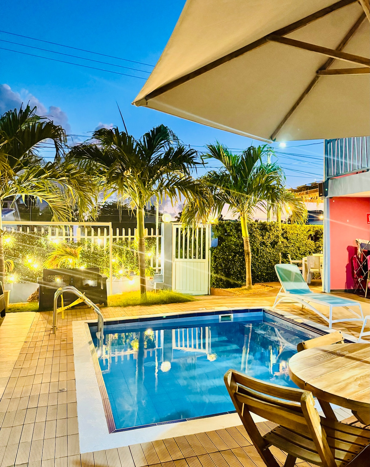 A tranquil outdoor area features a small pool surrounded by lush greenery and palm trees. Wooden lounge chairs are positioned nearby, with a shaded table and chairs offering a comfortable space to relax. Soft lighting enhances the inviting atmosphere during the evening.