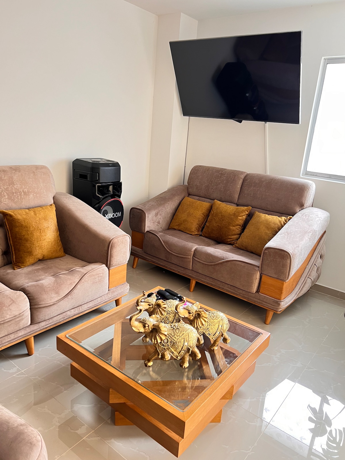 A light, modern living area features two cozy sofas adorned with gold accent pillows. A wooden coffee table sits in front, displaying a decorative piece shaped like animals. A wall-mounted TV is positioned above for entertainment, with natural light coming through a nearby window.