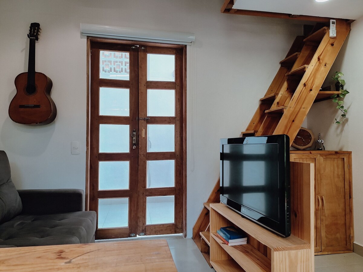 A cozy living area features a wooden door with glass panels, inviting natural light. A plush gray sofa is positioned toward a wooden TV stand, while a guitar hangs on the wall nearby. A wooden ladder leads to the mezzanine level above.
