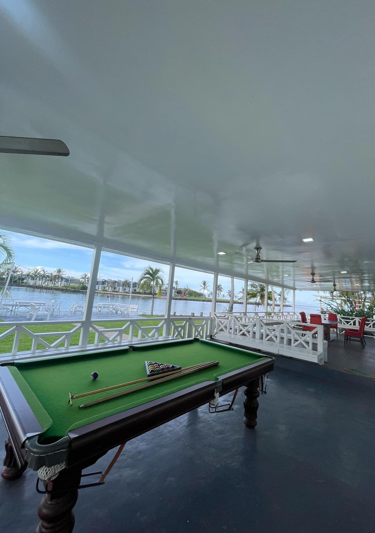 An outdoor patio area features a pool table with a green felt surface. Large windows provide views of the waterfront and surrounding palm trees. Chairs and tables are arranged for dining, with additional seating visible in the background.