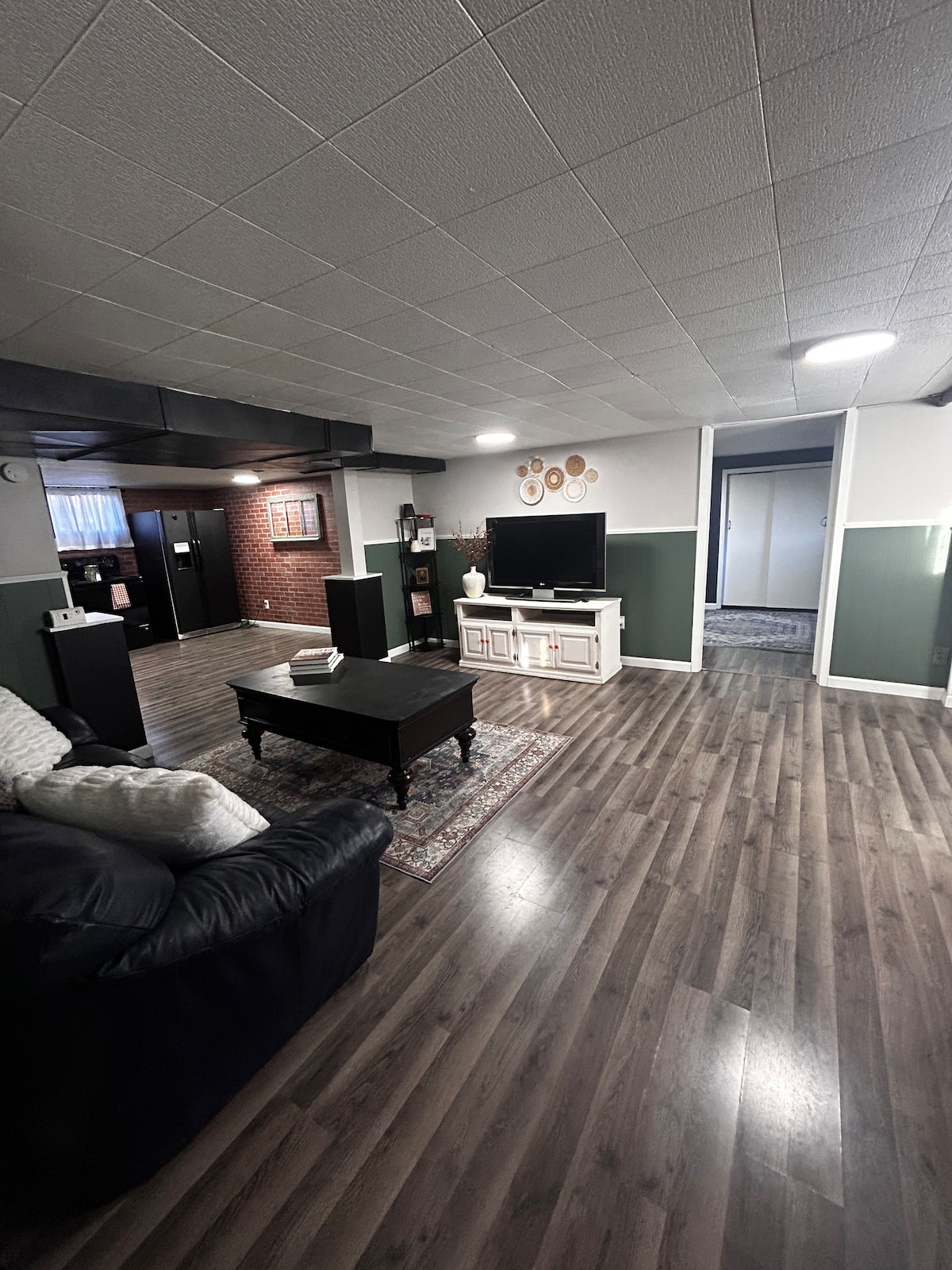 A spacious living area features comfortable seating arranged around a dark coffee table. A large TV is positioned on a white console against a wall. The room is complemented by wood-like flooring, modern decor, and a welcoming color scheme.