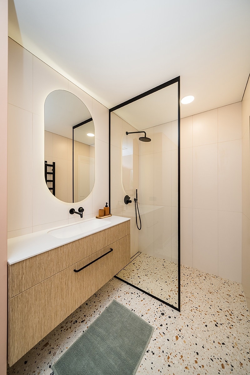 A modern bathroom features a walk-in shower enclosed by a glass panel. A minimalist vanity with a rounded mirror is complemented by natural wood and black fixtures. The floor showcases a textured surface with small colored stones, while a light green mat is positioned in front of the sink.