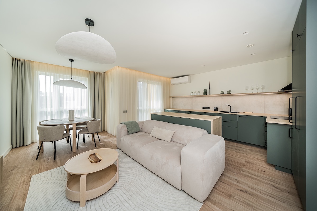 A modern living space features a light-coloured sectional sofa and a round coffee table. A dining area with a round table and two upholstered chairs is visible. The kitchen area includes green cabinetry and a neutral countertop, with large windows allowing natural light to fill the room.