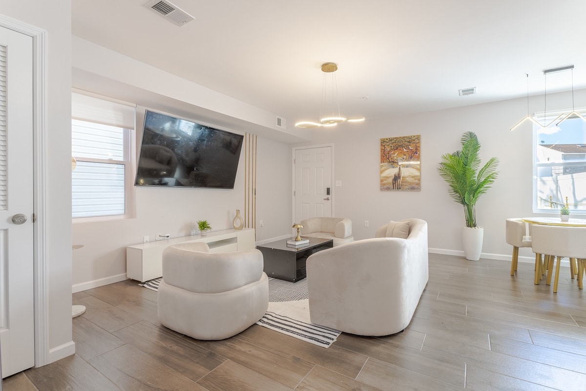 A spacious living room is featured, illuminated by natural light from a window. Two curved sofas face a mounted flat-screen television. A decorative plant adds a touch of greenery, while a dining area with a circular table is positioned in the background.