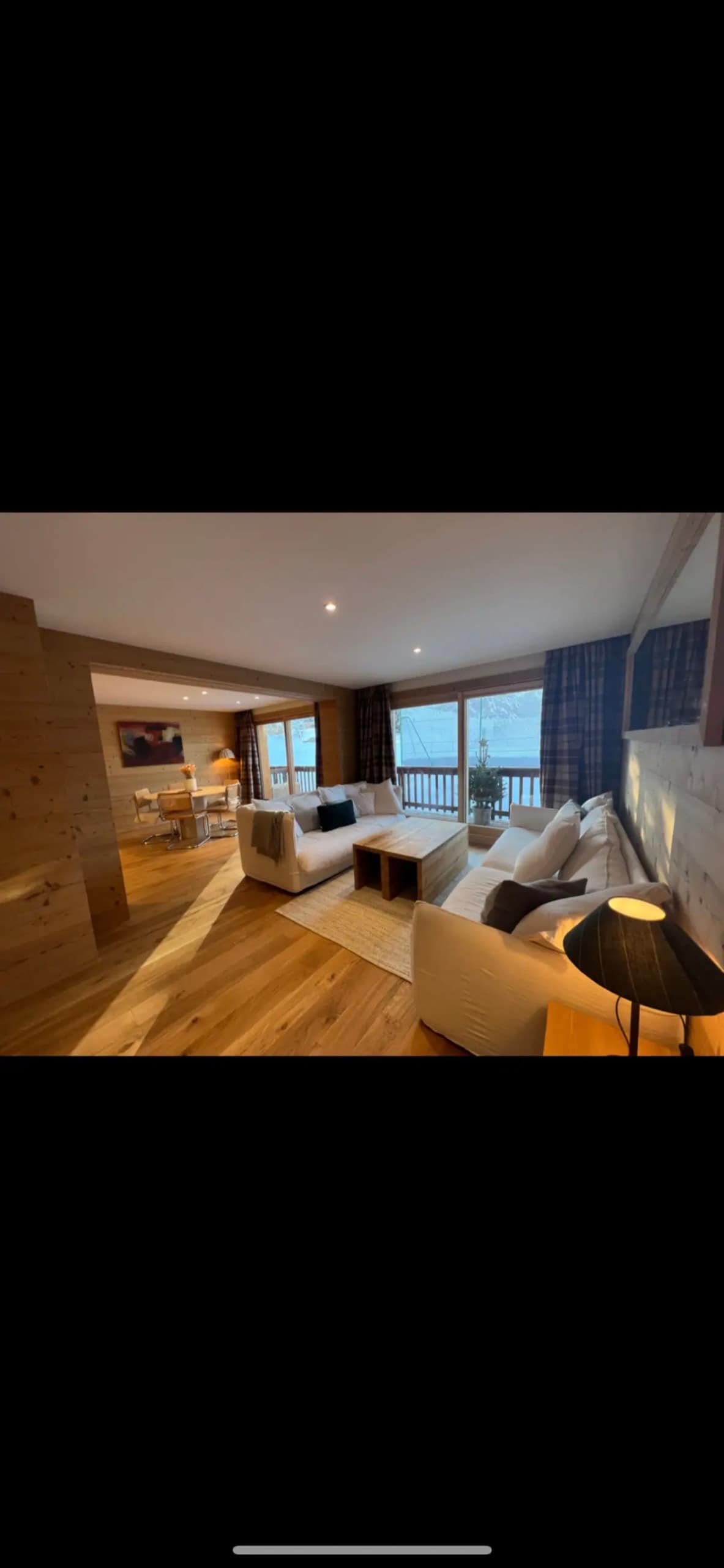 A spacious living area features cozy seating arranged around a wooden coffee table, with large windows allowing natural light to illuminate the room. Neutral-toned walls and wood accents create a warm aesthetic, while a lamp provides soft lighting.