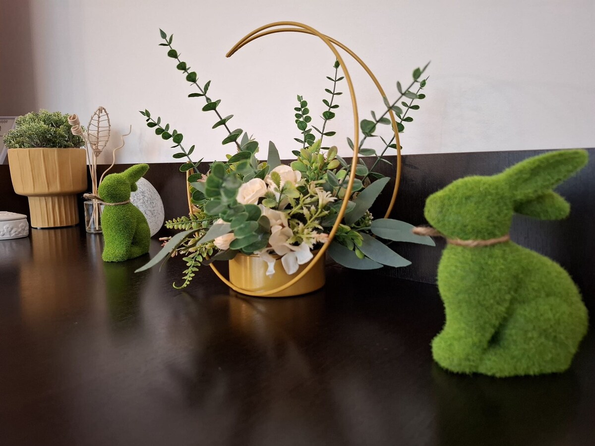A decorative arrangement is displayed on a dark wooden surface. A round golden holder contains greenery and neutral-colored flowers. Two green, moss-like rabbits are positioned on either side, adding a touch of nature to the decor.