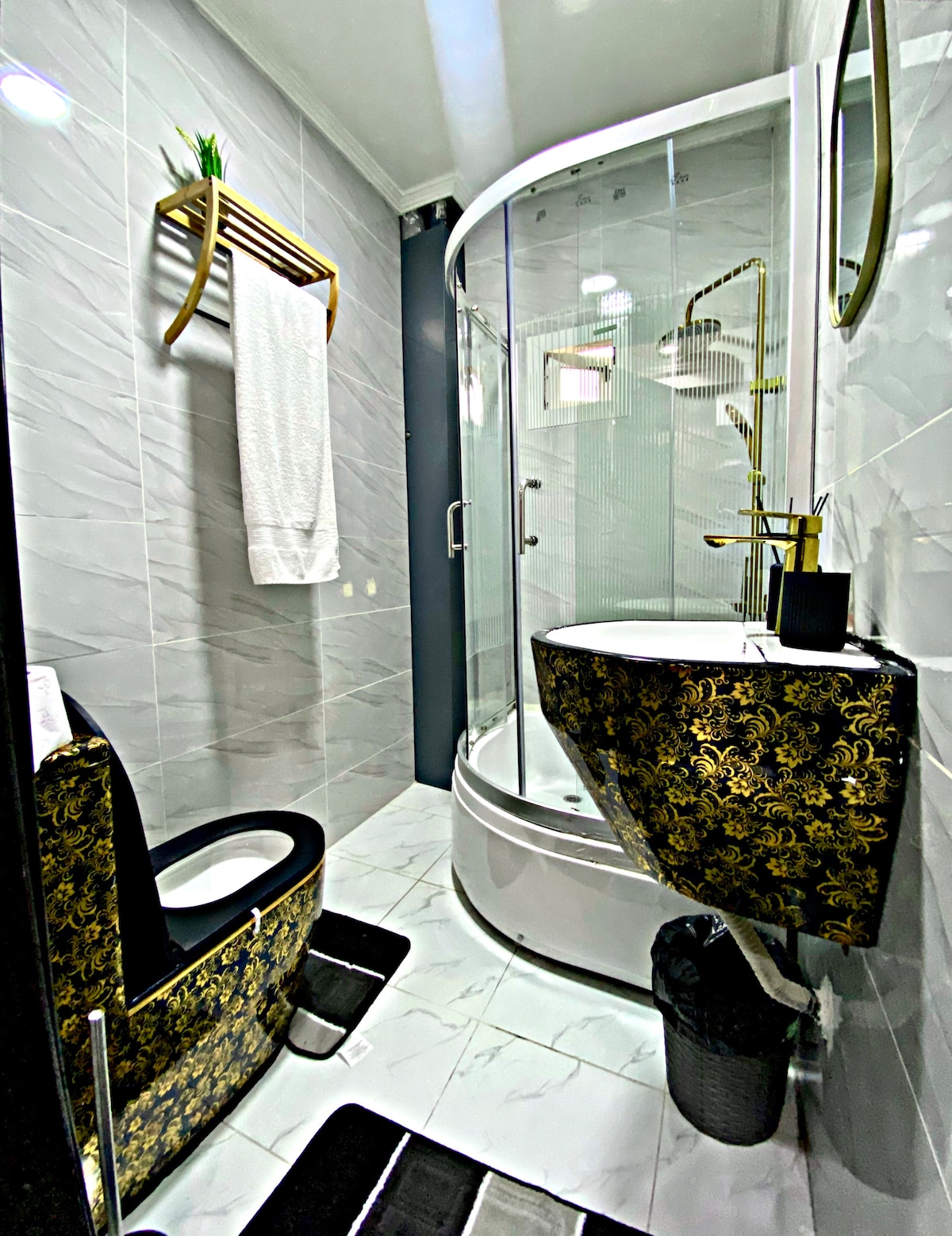 A modern bathroom is displayed with sleek gray tiles and a curved shower enclosure. A stylish toilet is outlined in a decorative black and gold pattern. A towel hangs on a wooden rack, and a round mirror reflects the interior lighting, enhancing the minimalist design.
