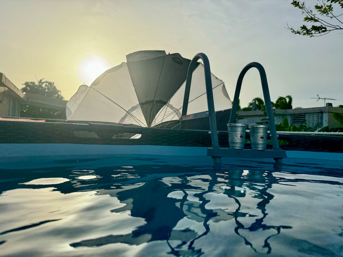 The tranquil swimming pool reflects the soft hues of sunrise, with a large umbrella positioned nearby. Two beverage holders are visible beside the pool's edge, contributing to a serene atmosphere. The background features lush greenery, enhancing the outdoor space.