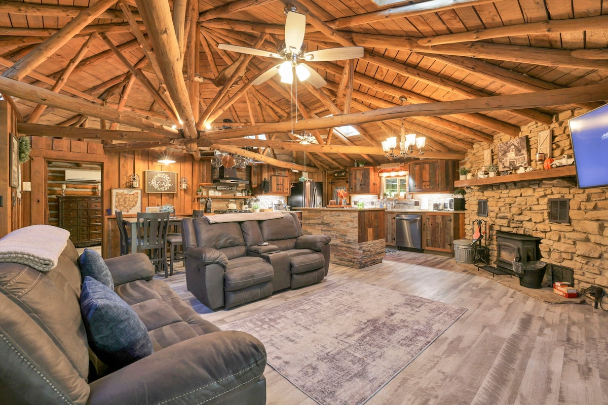 The open-concept living area features a cozy seating arrangement with plush sofas on a textured rug. The wooden ceiling showcases exposed beams, creating a rustic charm. A stone fireplace is positioned against the wall, while the kitchen is visible in the background, highlighting modern appliances.