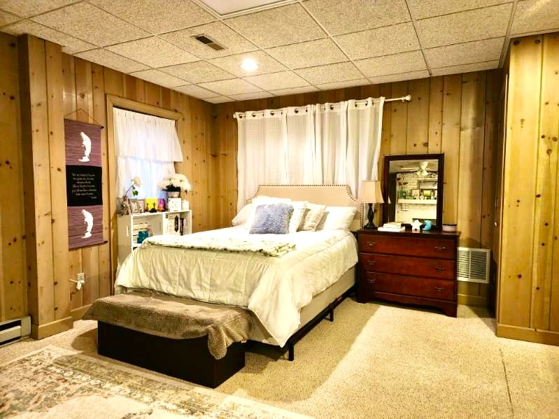 A spacious lower level bedroom features wooden paneling and soft carpet. A queen bed, adorned with layered bedding, is positioned against a wall with a window dressed in white curtains. A dresser and a mirrored stand complement the room, adding functionality.