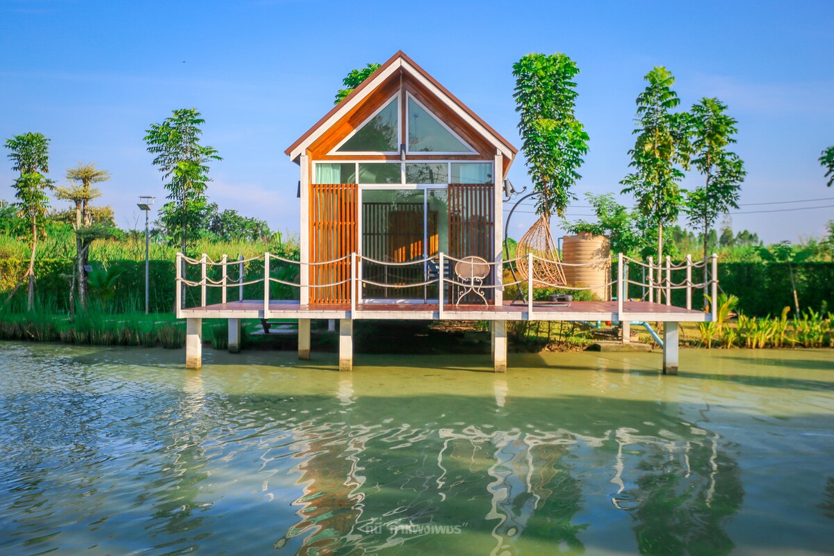 A charming riverside cottage is seen on stilts above the calm water. Large glass windows allow natural light to fill the interior, while a wooden deck provides a space to relax. Lush greenery surrounds the property, enhancing the serene environment.