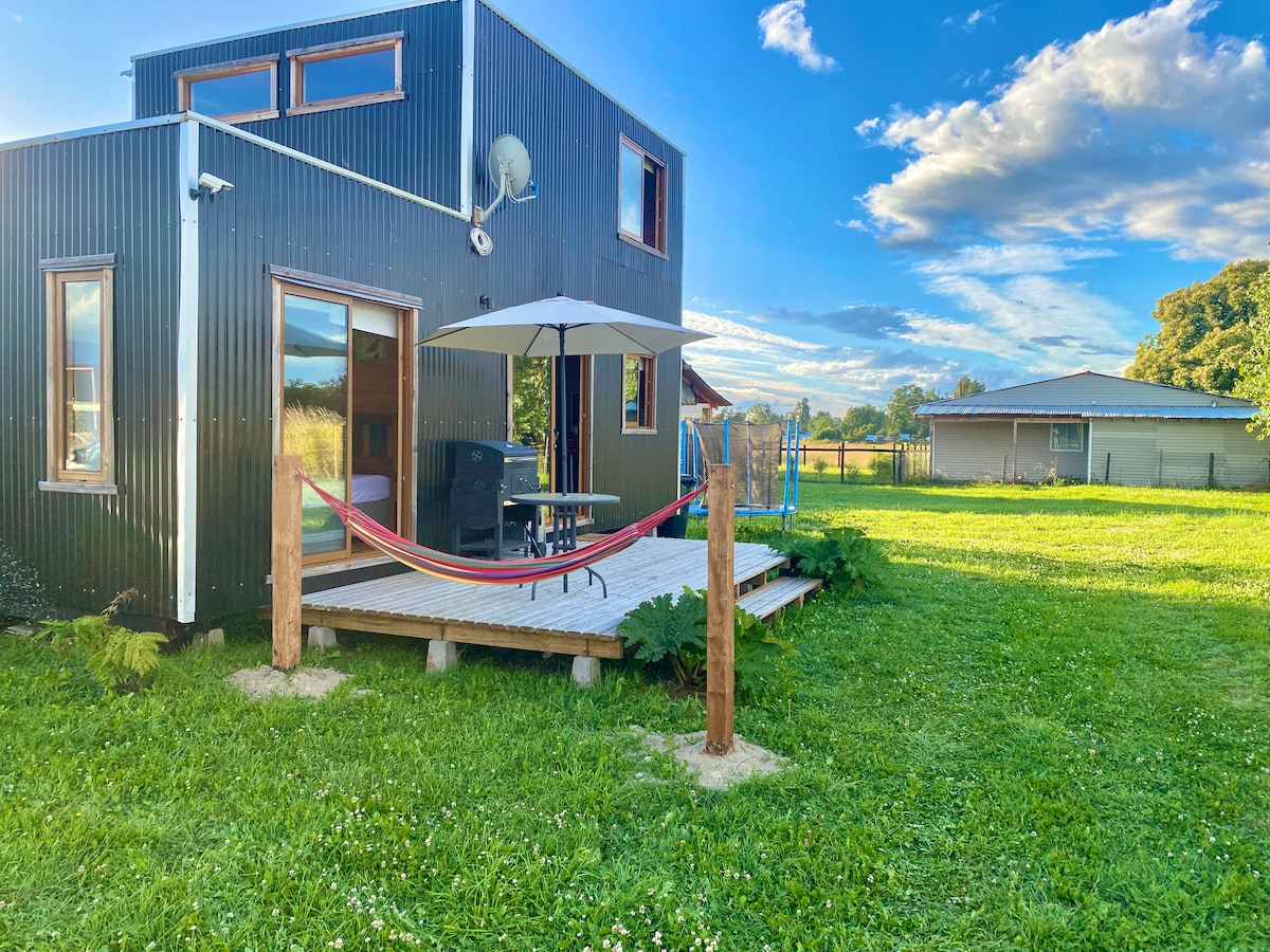 A modern black-clad structure is set against a vibrant green lawn. A wooden deck features a hammock and a table with chairs, complemented by a large umbrella for shade. The surrounding landscape includes trees and a distant building, all under a bright sky.