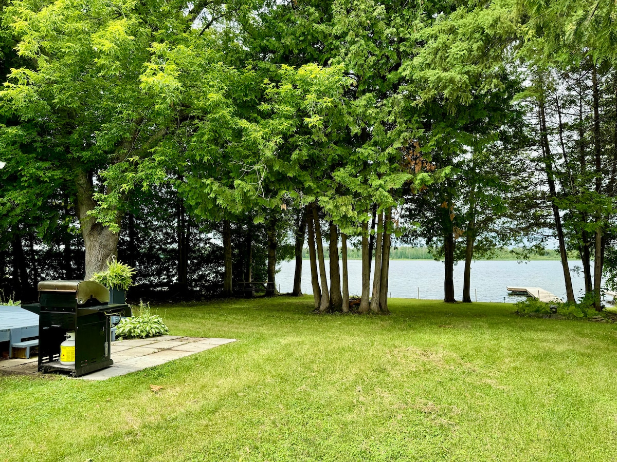 A serene outdoor space is framed by lush green trees, leading to a calm body of water. A flat stone patio features a grill, providing a perfect setting for outdoor cooking. The expansive yard is well-maintained, with ample green grass inviting relaxation.
