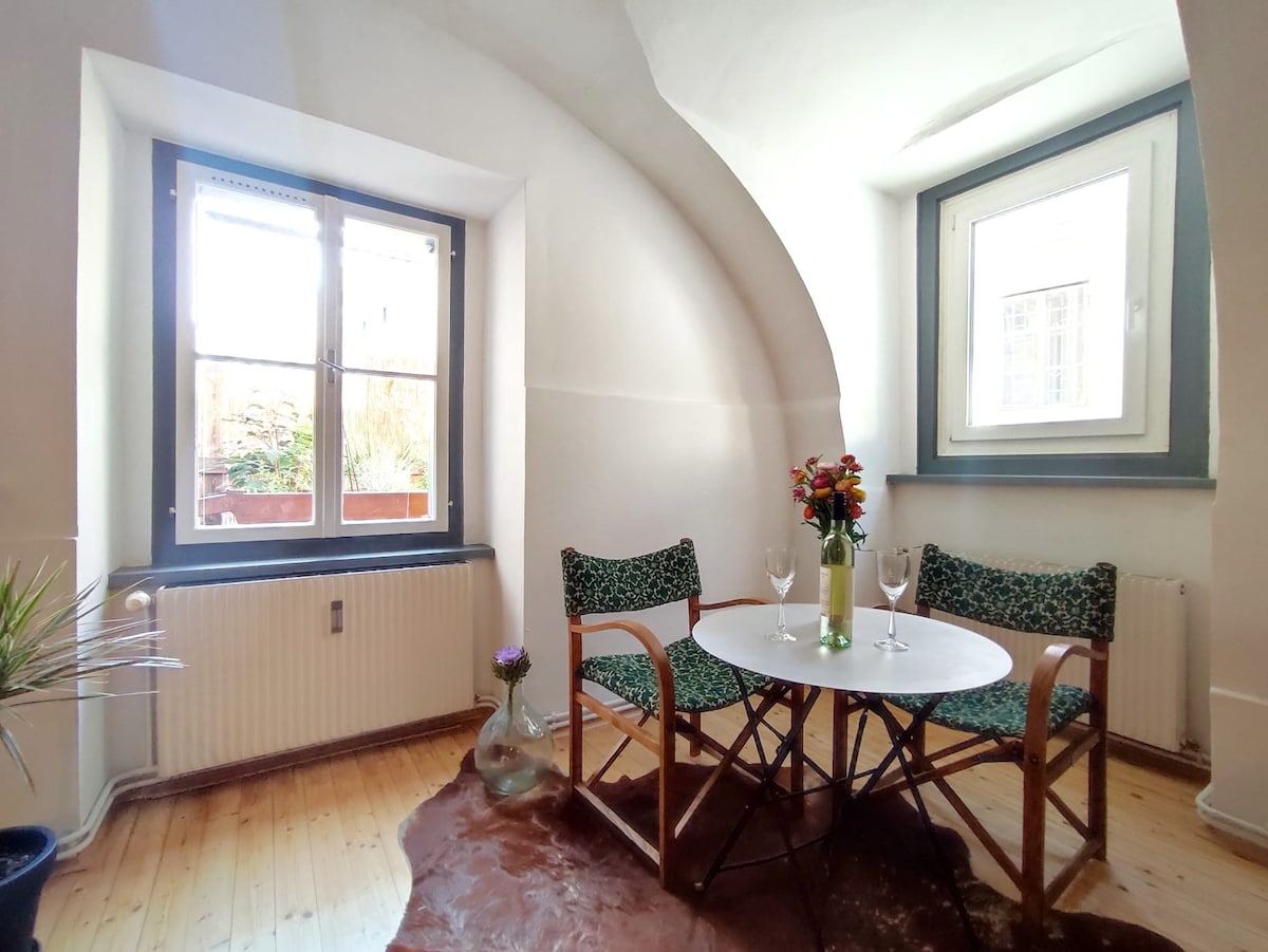 A cozy seating area is showcased with two patterned chairs beside a small round table. A vase filled with flowers sits in the center, and light enters through the large windows, highlighting the gentle curvature of the ceiling.