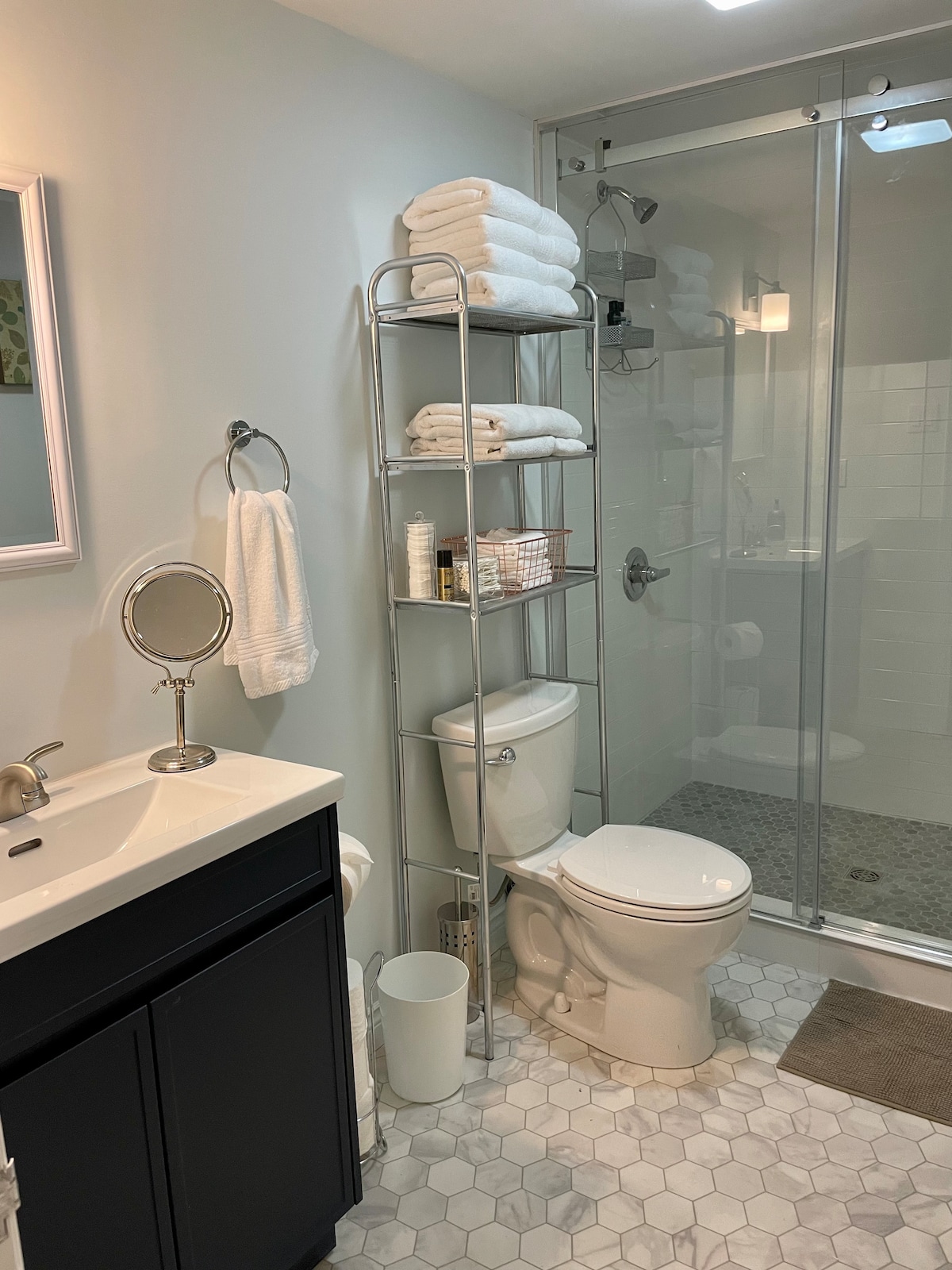 A modern bathroom is equipped with a glass-enclosed shower and a compact vanity with a sink. Fresh towels are neatly arranged on a metallic shelving unit. The space features light-colored walls, a large mirror, and a stylish tile floor that enhances the overall design.