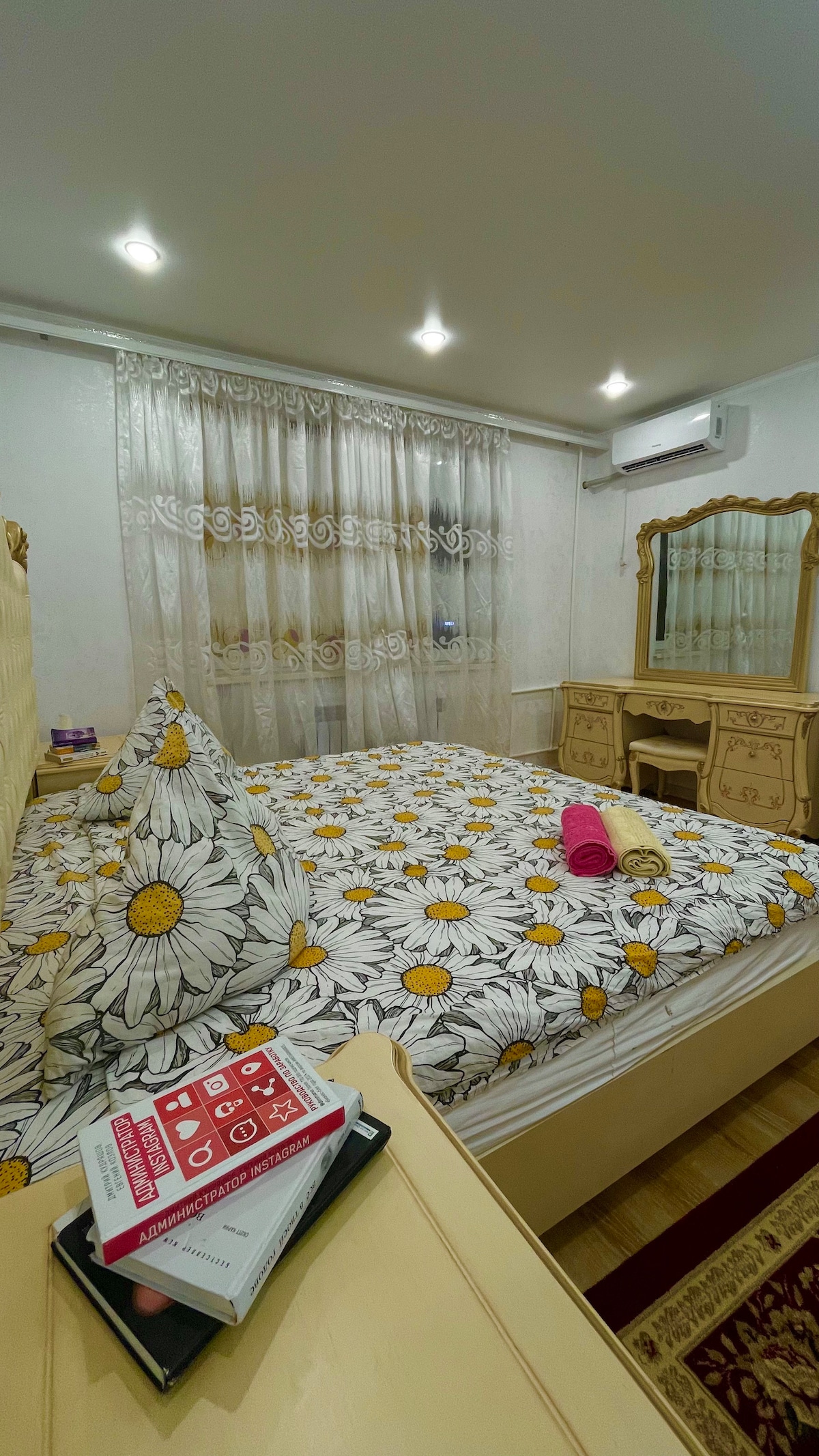 A bright bedroom features a spacious bed adorned with a floral-patterned duvet and large pillows. Soft lighting illuminates the space, complemented by sheer curtains that soften natural light. A dresser with a mirror and decorative items can be seen, creating an organized and inviting area.