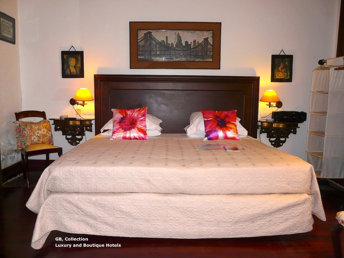 A spacious bed is presented with decorative pillows featuring bold floral patterns. A light-colored bedspread covers the bed, complemented by two bedside lamps providing soft illumination. Wall art depicting a city skyline enhances the room's character, with a wooden chair positioned nearby.