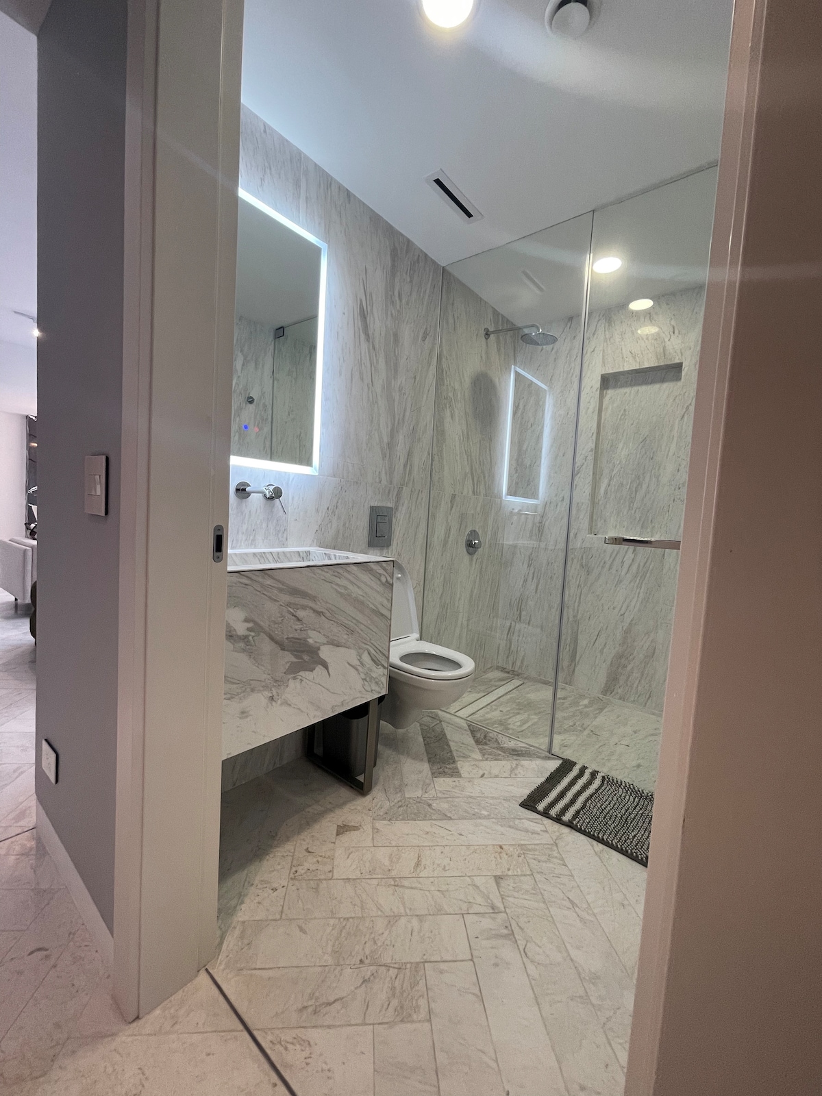 A modern bathroom is featured, showcasing sleek marble walls and floors. A glass shower enclosure is visible, complemented by contemporary fixtures. Soft lighting from a backlit mirror creates a calming ambiance. A striped mat lies on the floor for added comfort and style.