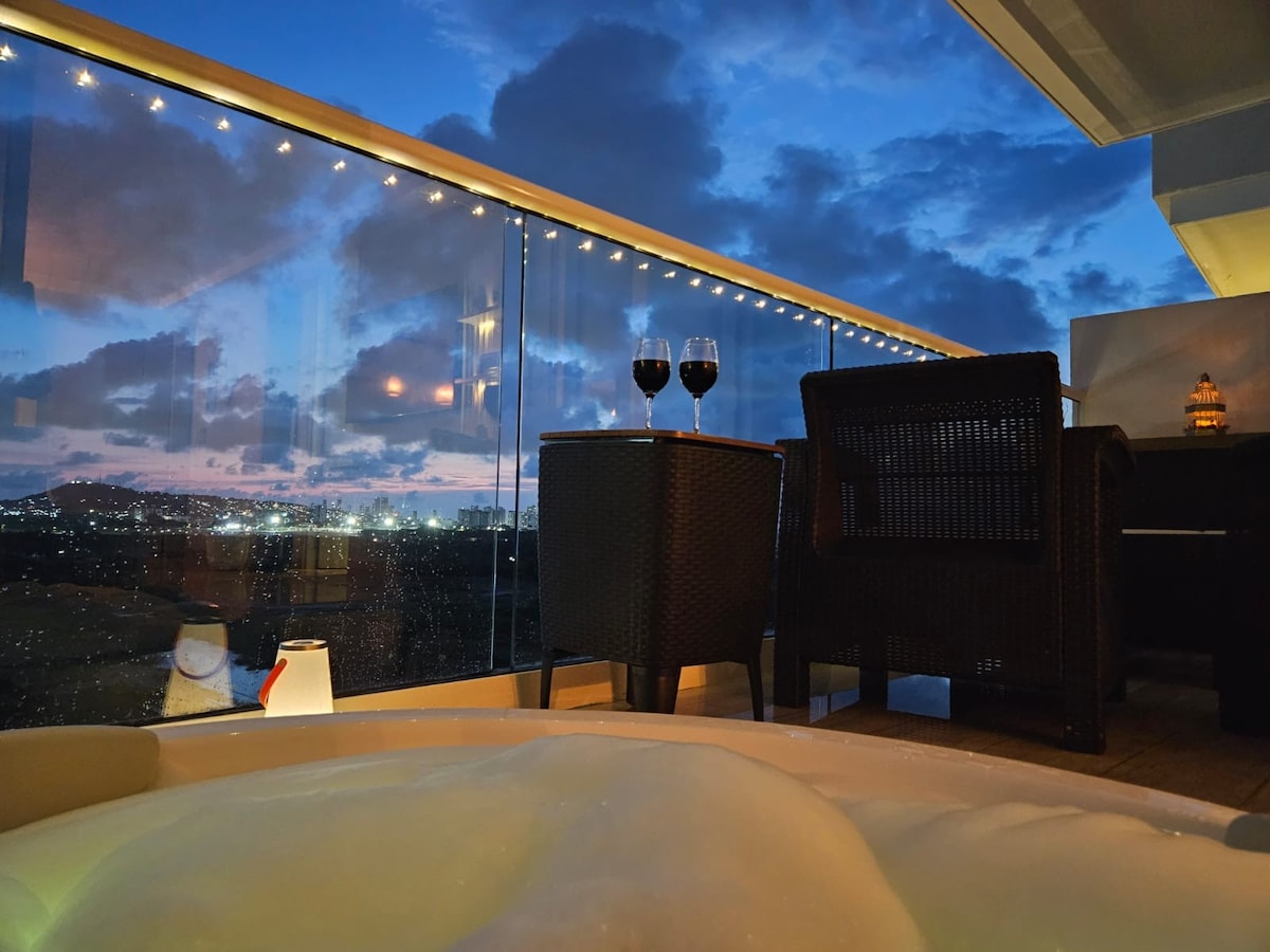 A balcony scene presents a relaxing setting with two glasses of red wine placed on a table. Comfortable seating is arranged beside a private Jacuzzi, with views of a twilight sky reflected in the glass railing. Soft lighting adds an inviting touch.